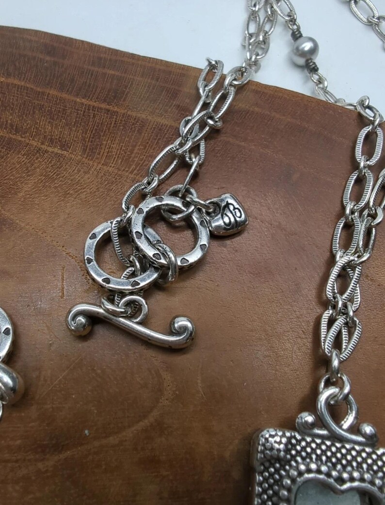 Peut inclure: Gros plan sur un collier argent&eacute; avec un fermoir &agrave; bascule. Le collier pr&eacute;sente divers maillons de cha&icirc;ne et &eacute;l&eacute;ments d&eacute;coratifs, dont un petit charme au design orn&eacute;. Le fond est une surface en bois.