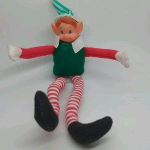 May include: A vintage elf doll with a green body, red arms, and striped red and white legs. The elf has a white collar, a green and white striped hat, and black shoes. The doll's arms are outstretched, and it is set against a white background.