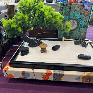 Puede incluir: Un jardín zen con marco negro, arena blanca, piedras negras, un pequeño puente de madera y una pagoda de madera en miniatura. El jardín está colocado sobre una base con estampado floral blanco y naranja.