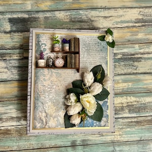 Puede incluir: Un álbum de recortes hecho a mano con un diseño floral azul y blanco. La portada presenta un estante de madera en miniatura con plantas en macetas, libros y un reloj.