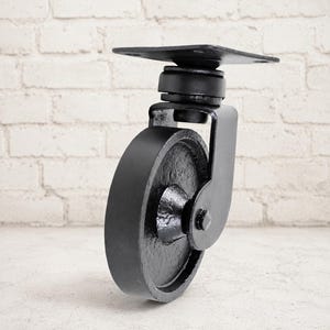 Puede incluir: Una rueda giratoria de metal negro de alta resistencia que cuenta con una placa de montaje plana con cuatro orificios pretaladrados, un soporte robusto y una rueda circular sólida con acabado texturizado para muebles industriales.