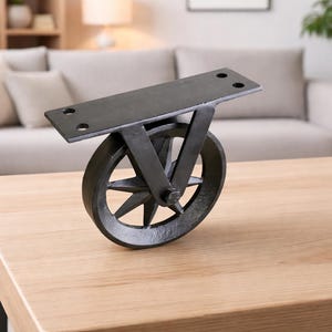 Puede incluir: Una rueda de metal negro con una placa de montaje rectangular, diseñada para muebles. La rueda tiene un diseño en forma de estrella y está montada sobre una superficie de madera clara. El fondo incluye un sofá y plantas.