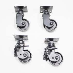 Puede incluir: Conjunto de cuatro ruedas giratorias de metal negro con una placa de montaje cuadrada. Dos de las ruedas tienen un freno de cadena.