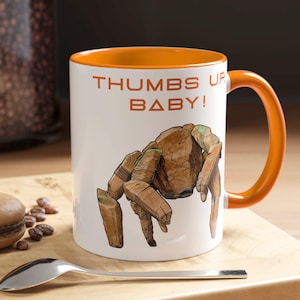 Peut inclure: Mug en céramique blanche avec intérieur et anse orange. L'illustration marron d'une créature et le texte "THUMBS UP BABY!" en orange ornent le mug. Une cuillère et un macaron sont sur une surface en bois.