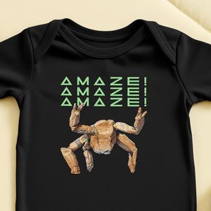 May include: Black baby bodysuit with a graphic of a crab with its claws raised and the word "AMAZE!" in green. The bodysuit is made of soft fabric and has short sleeves.