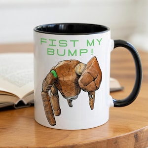 May include: White ceramic mug with a black interior and handle. The mug features a brown, robotic hand illustration with green accents and the text "FIST MY BUMP!" in green.