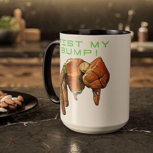 May include: White ceramic mug with a black handle and rim. The mug features a cartoon illustration of a brown fist bumping another brown fist. The text "FIST MY BUMP!" is printed in green above the illustration.