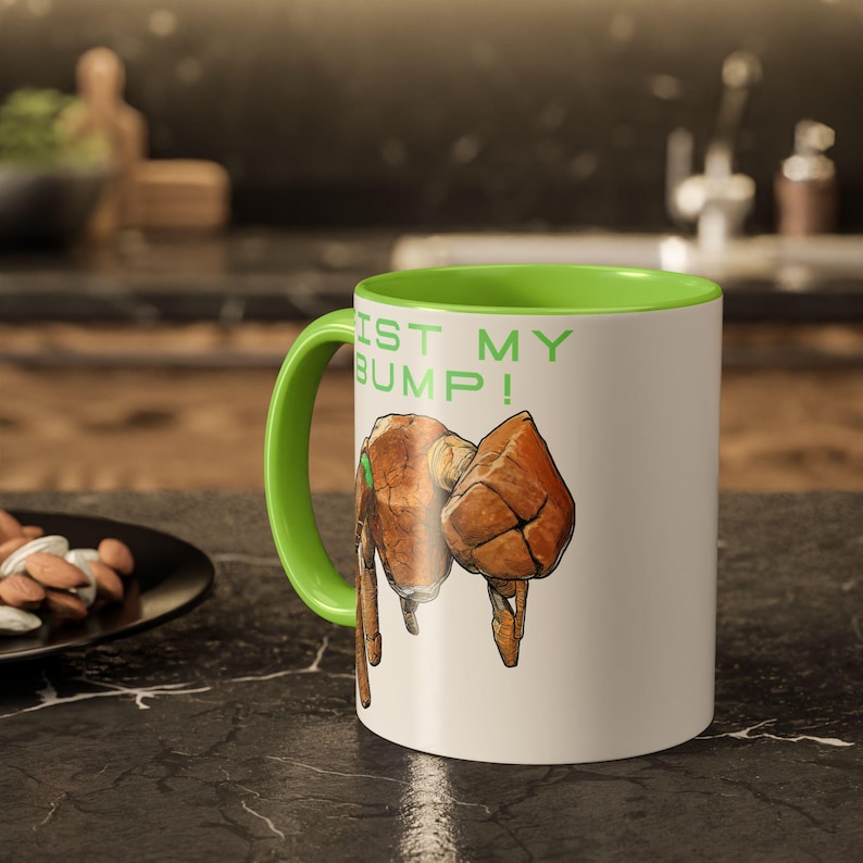 May include: White ceramic mug with a green interior and handle. The mug features a graphic of two brown fists and the text "FIST MY BUMP!" in green. The mug is sitting on a black surface.