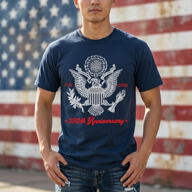 May include: Navy blue t-shirt featuring a white and red graphic of the American eagle, the text "1776" and "2026", and the words "250th Anniversary" in red. The shirt is worn in front of an American flag.