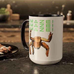 Puede incluir: Una taza de caf&eacute; blanca grande con asa e interior negros. La taza presenta una ilustraci&oacute;n de cangrejo estilizada marr&oacute;n con el texto "AMAZE!" en verde. La taza est&aacute; sobre una encimera oscura.
