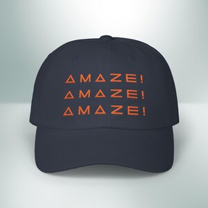 May include: Navy blue baseball cap with the word "AMAZE!" embroidered in orange. The text is repeated three times across the front of the cap. The cap has a curved brim and a rounded crown.