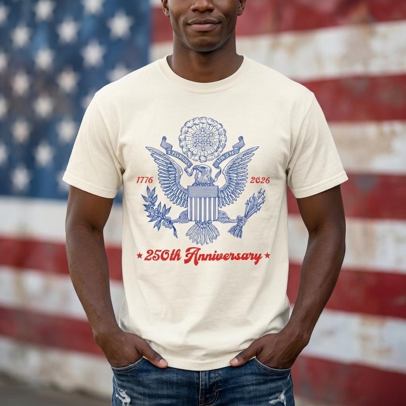 May include: Cream-colored t-shirt featuring a blue and red design commemorating the 250th anniversary. The design includes an eagle, the years 1776 and 2026, and the text "250th Anniversary". The background is an American flag.