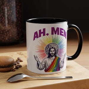 May include: White ceramic mug with a black interior and handle. The mug features a graphic of Jesus with a rainbow stole and a disco ball halo, with the words "AH, MEN" in purple. The mug is on a wooden surface.