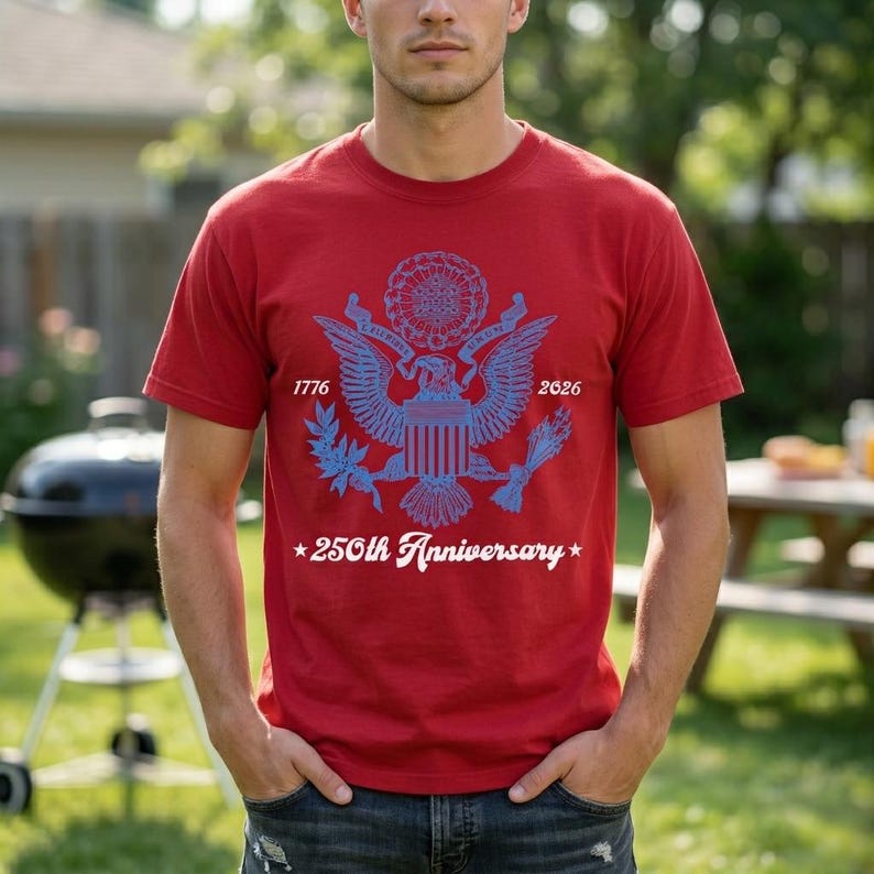 May include: A red t-shirt featuring a blue graphic of an eagle with the text "1776" and "2026". Below the eagle is the text "250th Anniversary" in white. The shirt is short-sleeved.