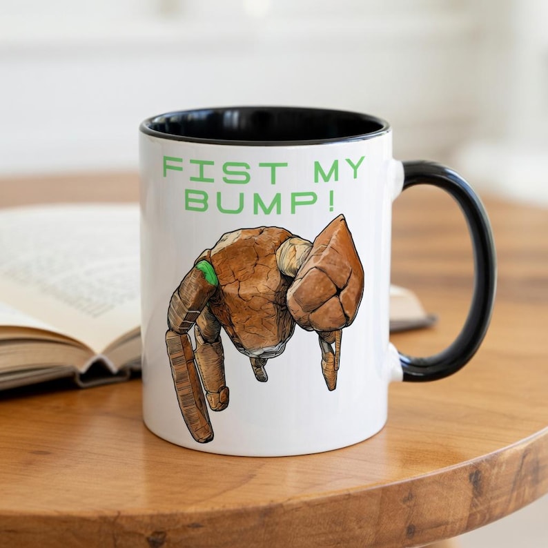 May include: White ceramic mug with a black interior and handle. The mug features a brown, robotic hand illustration with green accents and the text "FIST MY BUMP!" in green.
