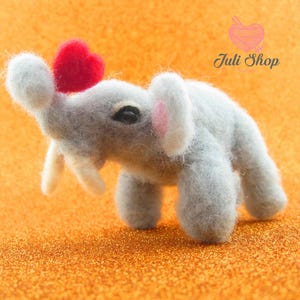 May include: A gray felt elephant figurine with a red heart on its head. The elephant has white tusks and pink-lined ears. The background is a glittery orange surface. The text "Juli Shop" is in the upper right corner.