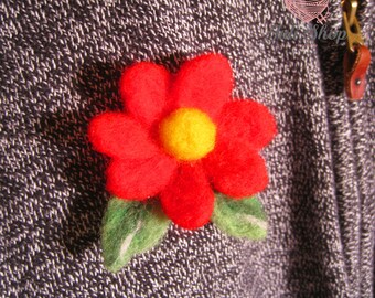 Könnte beinhalten: Eine rote Filzblume mit gelber Mitte und grünen Blättern. Die Blume ist an einem grauen Pullover befestigt.