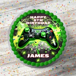 May include: A round birthday cake decorated with green icing and a video game controller design. The cake has the text "HAPPY 8TH BIRTHDAY JAMES" in white. The controller is black with green detailing.