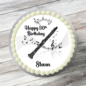 May include: A round cake with white icing and a decorative edge. The cake has a black and silver clarinet, musical notes, and the text "Happy 30th Birthday Steven."