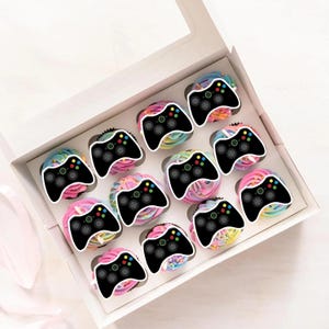 May include: A box of twelve cupcakes, each decorated with a black video game controller. The cupcakes have colourful rainbow frosting in shades of pink, blue, yellow, and green.
