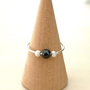 May include: A thin silver ring featuring a single round, polished black bead at the centre, flanked by two smaller, vertically ribbed silver beads. The ring is displayed on a smooth, light-coloured wooden cone stand, highlighting the contrast between the metallic silver band and the dark, glossy central bead.