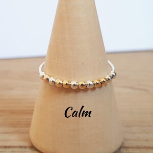 Könnte beinhalten: Ein zarter Ring mit abwechselnd goldenen und silbernen Perlen auf einem weißen Band. Das Wort "Calm" steht in schwarzer Schrift darunter. Der Ring wird auf einem Holzkegel präsentiert.