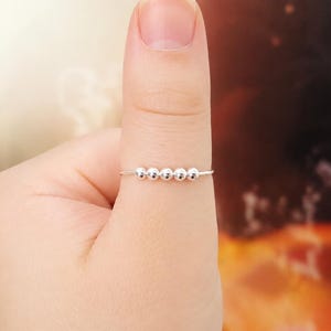 Puede incluir: Un anillo fino de plata con cinco pequeñas cuentas plateadas, lisas y redondas, alineadas horizontalmente en el centro de la banda. Las cuentas están diseñadas para deslizarse a lo largo de la fina banda de alambre que se lleva en el pulgar.