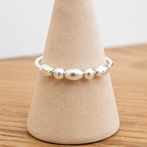 May include: A silver ring featuring a series of five polished, smooth metal beads arranged in a row along the front of the band. The beads alternate in size, with three oval-shaped beads and two smaller spherical beads positioned between them.