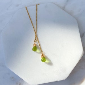Puede incluir: Un collar de cadena dorada con dos colgantes de piedra verde. Las piedras preciosas están envueltas en alambre dorado y tienen una forma de lágrima facetada.