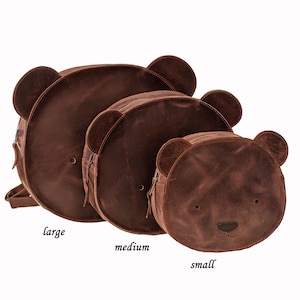 May include: Three brown leather bear-shaped bags in varying sizes: large, medium, and small. Each bag features a circular design with bear ears, a zipper closure, and embroidered facial details. The bags are arranged in a row, showcasing their size differences.