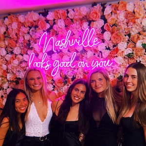 May include: Five friends pose in front of a pink flower wall with a neon sign that reads "Nashville looks good on you".