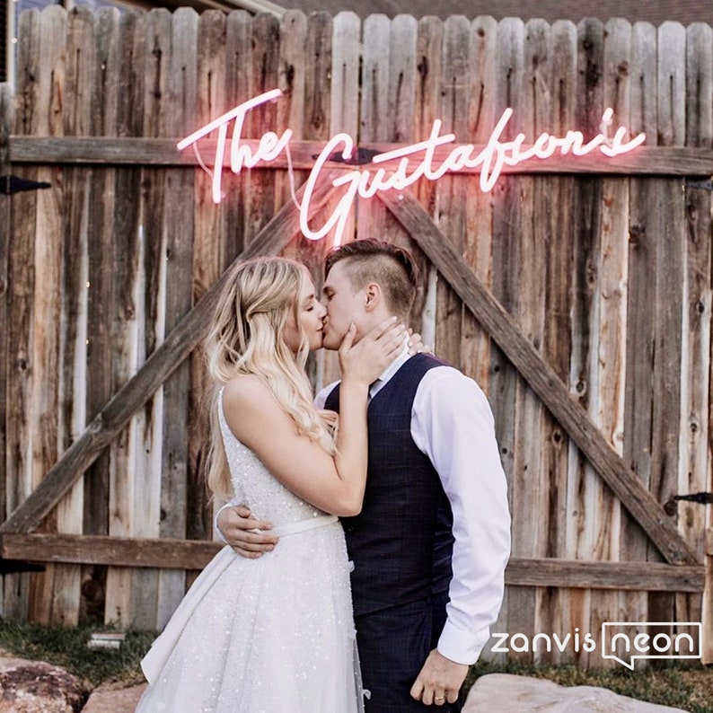 May include: A pink neon sign displaying "The Gustafson's" in cursive script is mounted on a wooden fence. A couple is kissing in front of the sign. The image is taken outdoors, with a logo in the bottom right corner.