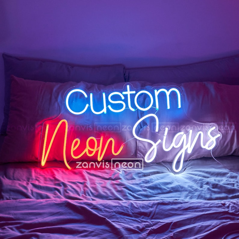 May include: Custom neon signs with the words "Custom Neon Signs" in blue, red, and white neon lights. The sign is on a bed with white and pink bedding.