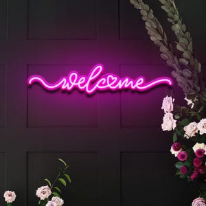 May include: A neon sign in a cursive font that spells "welcome" in bright pink. The sign is mounted on a dark wall, with floral arrangements in the background. The word "welcome" has a heart shape.
