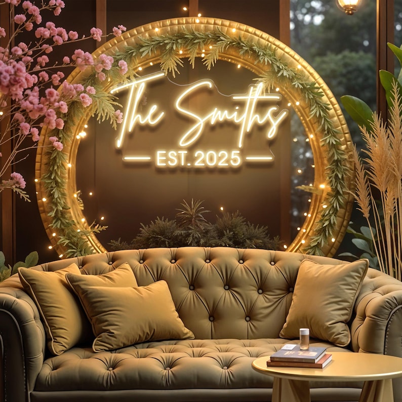 May include: A gold-framed sign with the text "The Smiths - EST. 2025" in white neon. The sign is decorated with foliage and fairy lights. Beneath, an olive green, button-backed sofa with matching cushions creates a warm, inviting atmosphere.