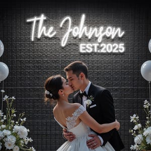 May include: A couple kissing beneath a neon sign reading "The Johnson EST. 2025". The bride is in a white wedding dress, and the groom wears a black tuxedo. White balloons and floral arrangements are on either side of the couple.