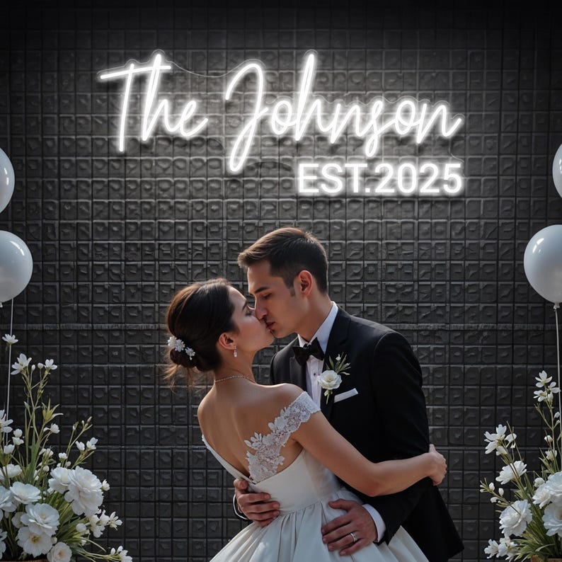 May include: A couple kissing beneath a neon sign reading "The Johnson EST. 2025". The bride is in a white wedding dress, and the groom wears a black tuxedo. White balloons and floral arrangements are on either side of the couple.