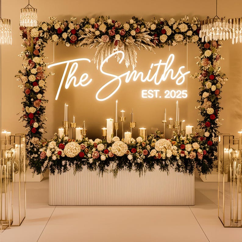 May include: A wedding reception table adorned with floral arrangements, candles, and a neon sign reading "The Smiths EST. 2025". The white table has a textured front. The flowers are red, white, and cream-colored, creating an elegant display.