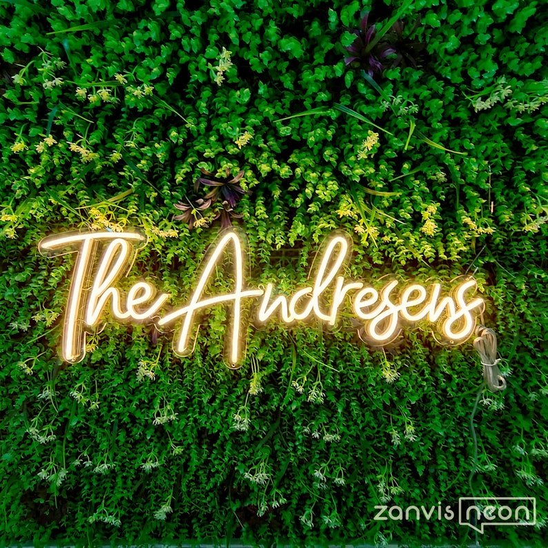 May include: A neon sign displaying "The Andresens" in a cursive script, glowing with a warm, golden light. The sign is set against a backdrop of vibrant green foliage. The "zanvis neon" logo is visible in the bottom right corner.