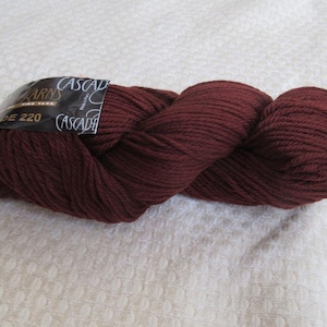 Op de afbeelding: Een streng Cascade-garen in een diepe bordeauxrode kleur. Het garen is strak gewikkeld en heeft een zachte, getextureerde uitstraling. Het etiket vermeldt "Cascade Yarns" en "DE 220". Ideaal voor brei- en haakprojecten.