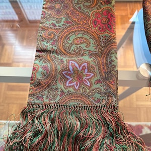 May include: A long rectangular scarf featuring a detailed paisley pattern in shades of olive green, orange, red, and light blue. The fabric has a smooth finish and is finished at the bottom edge with a dense fringe made of green and red threads.