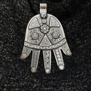 Könnte beinhalten: Ein silberner handförmiger Anhänger mit aufwendigen gravierten Designs. Der Anhänger zeigt ein zentrales Blumenmotiv und geometrische Muster.