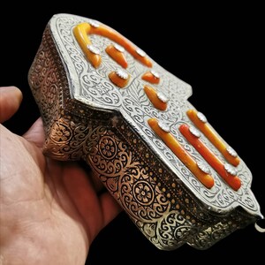 Peut inclure: Une boîte en forme de main de Hamsa en argent avec des détails gravés complexes et des accents en émail orange et blanc.
