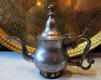 Op de afbeelding: Een zilveren theepot met een decoratief handvat en een gouden basis. De theepot staat op een wit oppervlak.