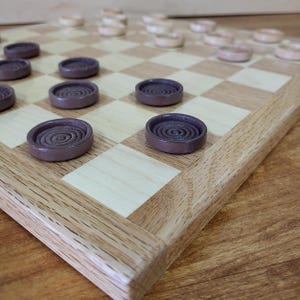 May include: A wooden checkerboard with a light wood and dark wood checkerboard pattern. The board has a light wood border. Dark brown checkers are placed on the board, with light brown checkers on the opposite side. The checkers have a spiral design.