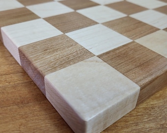 May include: A close-up of a wooden chessboard with alternating light and dark squares. The board is made of natural wood, with a light-colored wood and a darker, reddish-brown wood. The squares are precisely aligned, creating a classic checkerboard pattern.