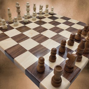 May include: A wooden chessboard with dark brown and light wood squares. Chess pieces are arranged on the board, with light-colored pieces on one side and dark brown pieces on the other. The board is set up for a game of chess.