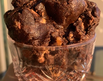 May include: A close-up of a glass bowl filled with a large amount of dark brown chocolate cookie dough. The dough is soft and appears to be made with chocolate chips and nuts.