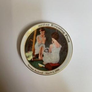 Peut inclure: Une assiette en c&eacute;ramique blanche avec un bord dor&eacute; pr&eacute;sente un tableau de Norman Rockwell repr&eacute;sentant une jeune femme se regardant dans un miroir. L'assiette porte l'inscription "The Saturday Evening Post" et "Norman Rockwell - March 8, 1954".