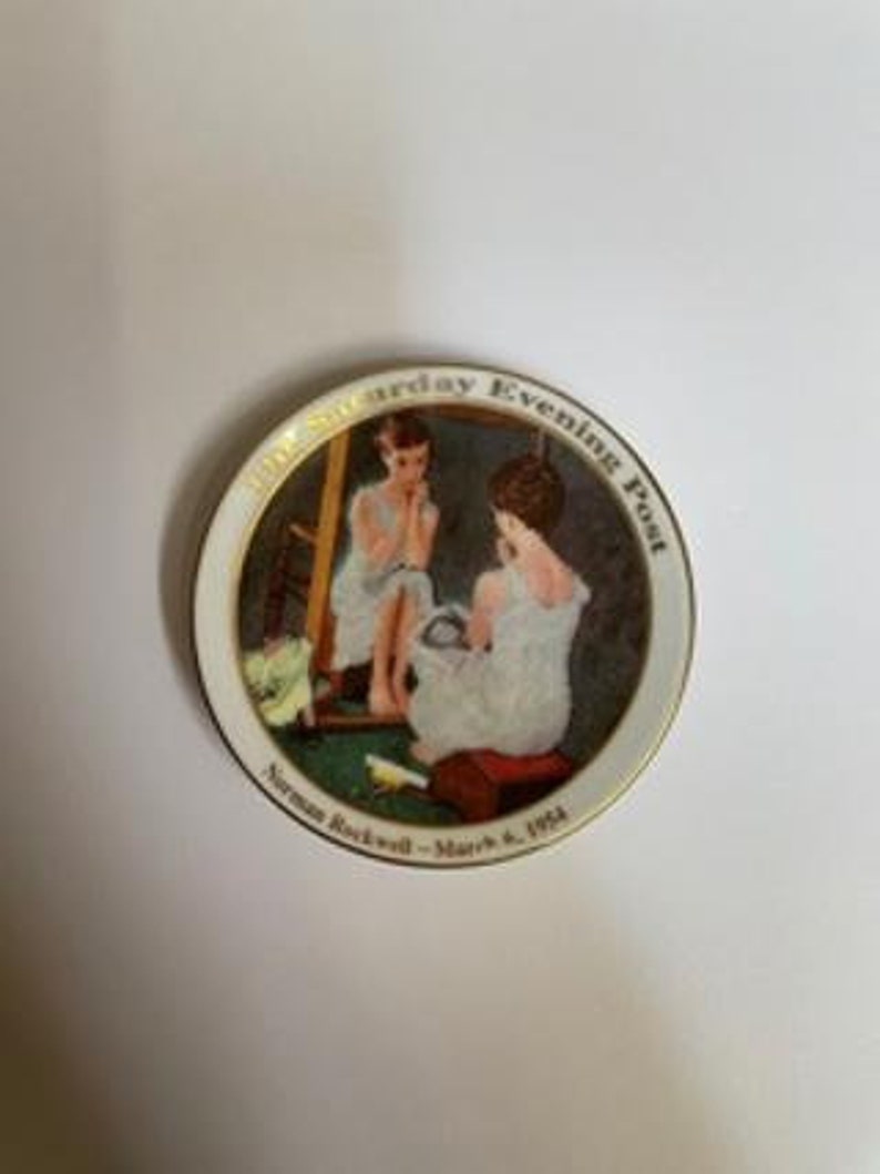 Peut inclure: Une assiette en c&eacute;ramique blanche avec un bord dor&eacute; pr&eacute;sente un tableau de Norman Rockwell repr&eacute;sentant une jeune femme se regardant dans un miroir. L'assiette porte l'inscription "The Saturday Evening Post" et "Norman Rockwell - March 8, 1954".